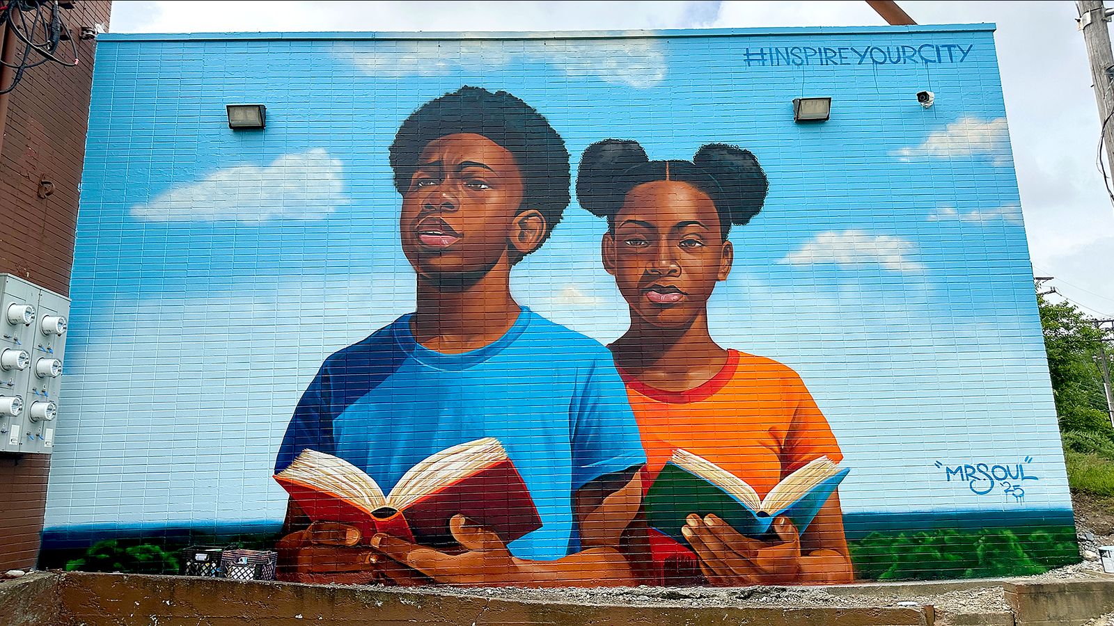 Inspire Buckeye-3 mural by mr.soul: a boy in a blue shirt and a girl in an orange shirt hold open books against a painted blue sky, with #INSPIREYOURCITY lettered at the top